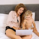 a mother and her young child sitting on a bed, engaged with a laptop, showcasing parenting and technology.