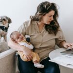 mother balancing work from home with parenting, holding baby and typing on laptop.