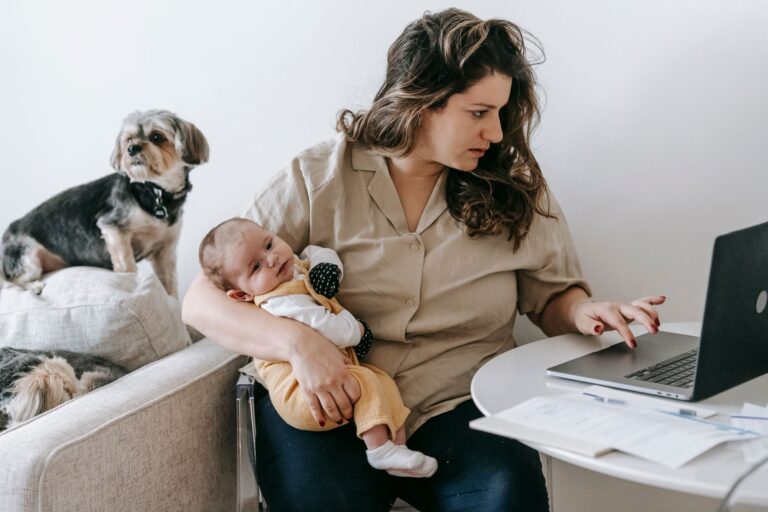 mother balancing work from home with parenting, holding baby and typing on laptop.