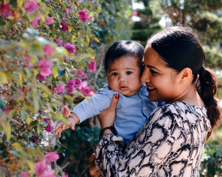 cheerful young ethnic female hugging cute baby boy and smiling while spending time together in garden near flowers