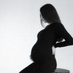 monochrome side view portrait of a pregnant woman sitting on a stool, embracing baby bump.