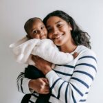 delighted african american mother cuddling cute little black baby while smiling and looking at camera on white backgroun