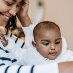 crop positive ethnic mother in casual clothes sitting with baby and smiling at home in daytime