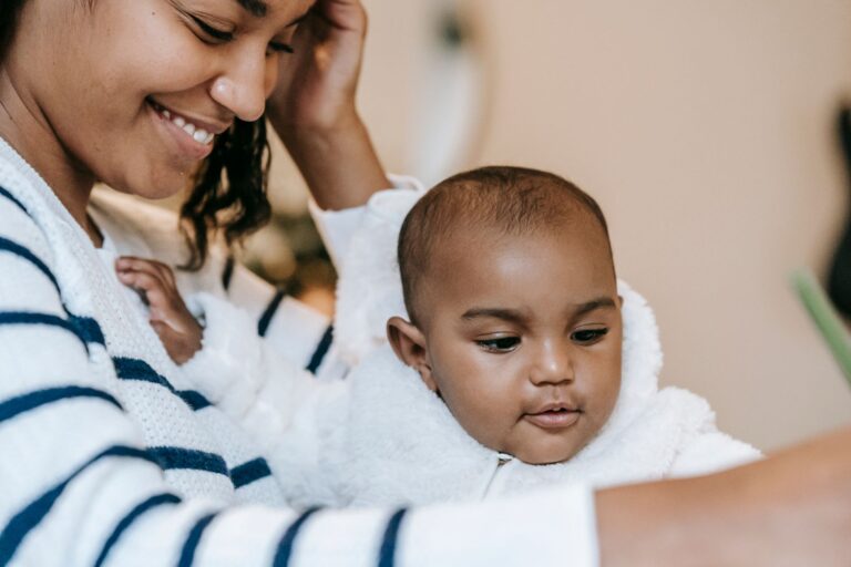 crop positive ethnic mother in casual clothes sitting with baby and smiling at home in daytime