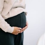 close-up of a pregnant woman in black pants and beige top holding her belly.