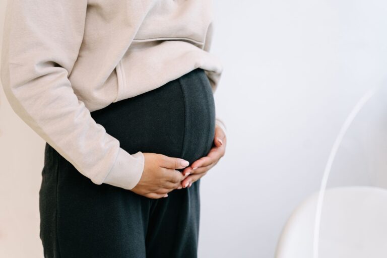 close-up of a pregnant woman in black pants and beige top holding her belly.