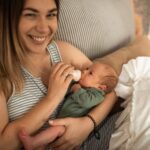 a mother lovingly feeds her newborn with a bottle while smiling at the camera.