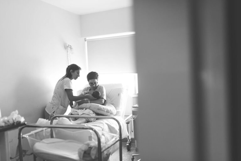 black and white image of a nurse assisting a mother with her newborn in a hospital room.