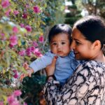 cheerful young ethnic female hugging cute baby boy and smiling while spending time together in garden near flowers