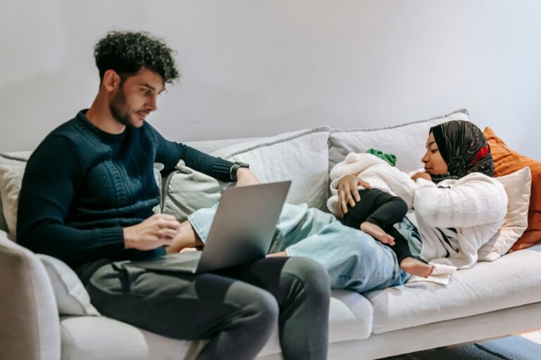 focused dad using netbook sitting near muslim mom feeding baby on sofa in living room
