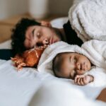 multiethnic father and newborn baby lying and sleeping together on comfortable bed near crop unrecognizable woman in day