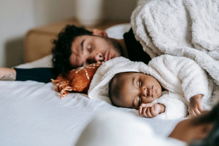 multiethnic father and newborn baby lying and sleeping together on comfortable bed near crop unrecognizable woman in day