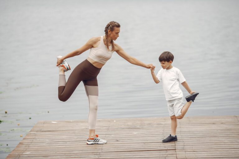 a mother and son holding a yoga pose together on a wooden pier by the water, promoting family fitness.