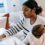 ethnic baby on laps hugging female in casual outfit with netbook at table while sitting in chair at light apartment near
