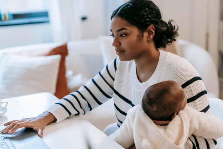 ethnic baby on laps hugging female in casual outfit with netbook at table while sitting in chair at light apartment near