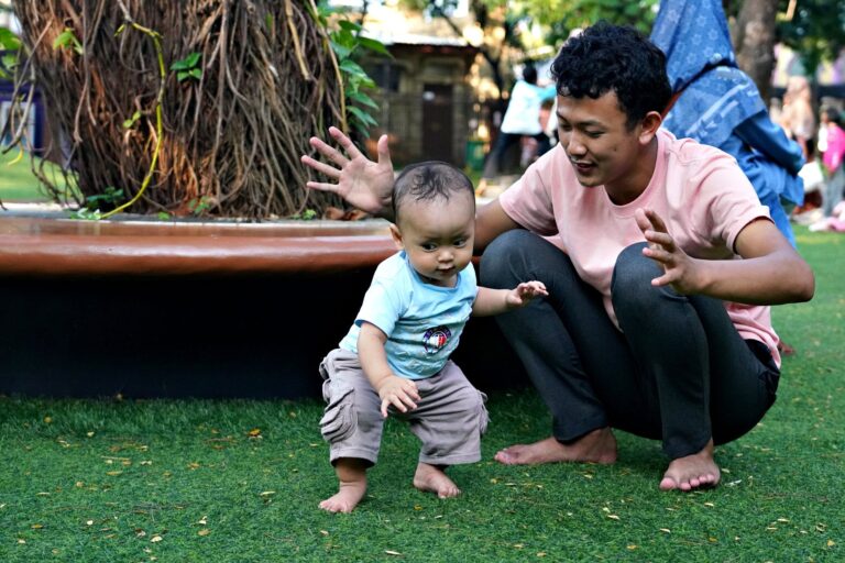 a father encourages his baby to take first steps on a sunny day in a park, barefoot on green grass.
