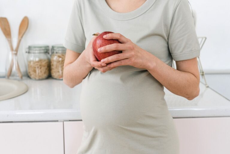 unrecognizable pregnant female with ripe red apple for healthy diet in hands standing near counter in light kitchen at h