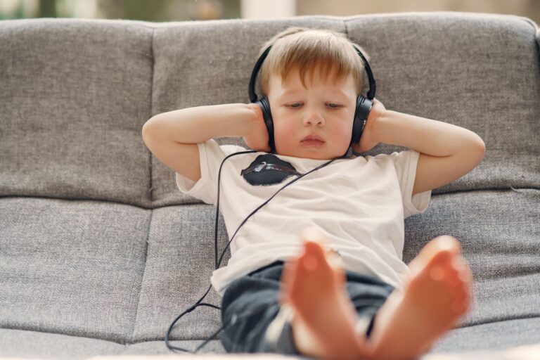 little boy in headphones attentively listening to music while sitting with barefoot on sofa in living room