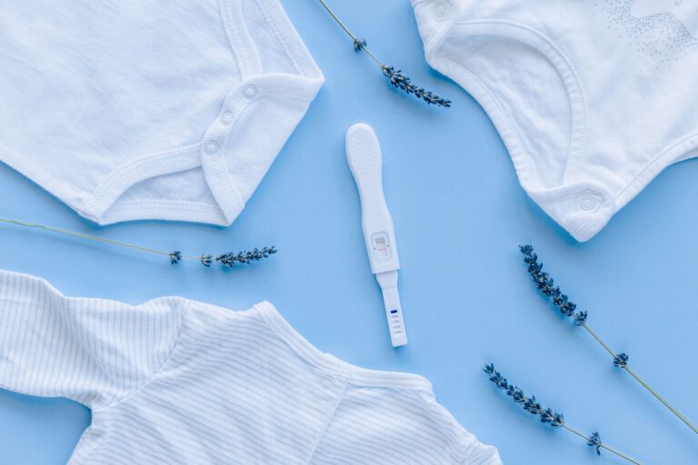 flat lay of baby clothes and positive pregnancy test on blue background.