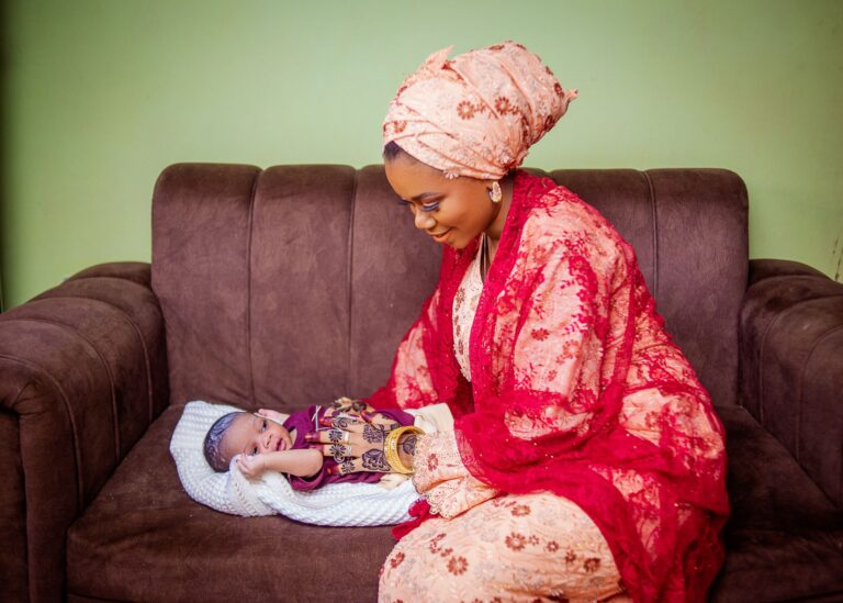 a mother in vibrant african attire lovingly gazes at her baby resting on a sofa.