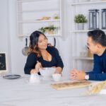 asian couple in a kitchen setting with a pregnancy announcement sign 'baby on the way', smiling at each other.
