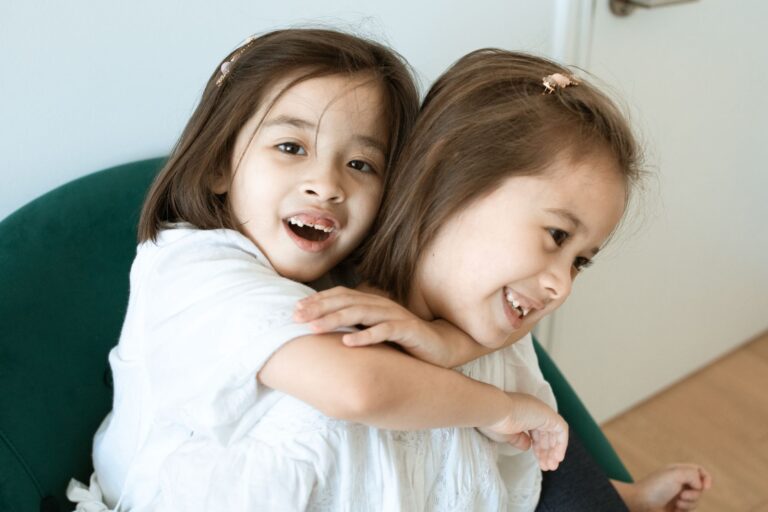 two smiling young girls hugging warmly on a chair inside a cozy room.