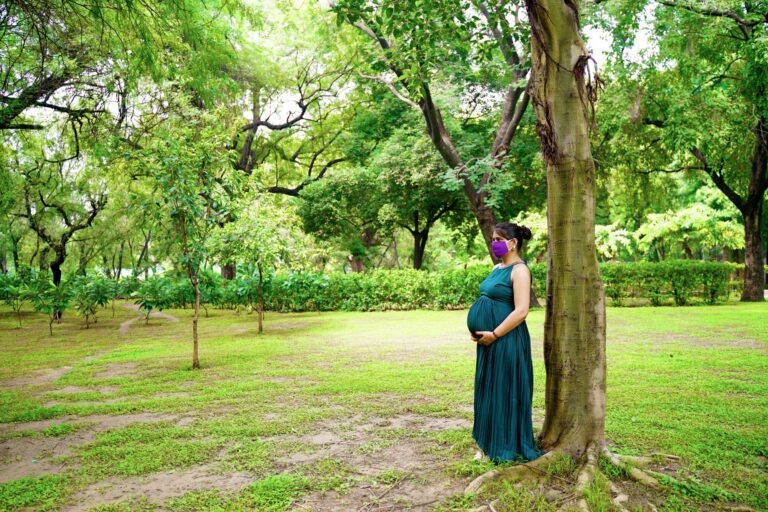 pregnant woman wearing a mask standing by a tree in a lush delhi park amidst pandemic.