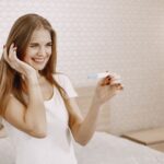 happy woman in white shirt holds positive pregnancy test, expressing joy indoors.
