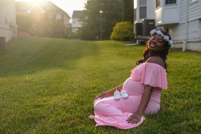 expecting mother sitting on lawn with baby shoes, wearing a flower crown.