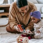 crop african american mother demonstrating book to little black daughter in headwear while sitting on rug with toy at ho