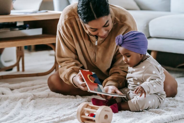 crop african american mother demonstrating book to little black daughter in headwear while sitting on rug with toy at ho