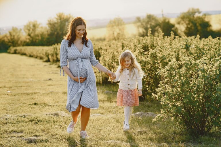 a pregnant mother holds her daughter's hand while walking in a sunny field surrounded by greenery.