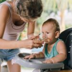 a mother helps her baby drink water from a glass while sitting in a high chair.