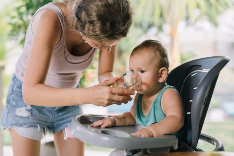 a mother helps her baby drink water from a glass while sitting in a high chair.