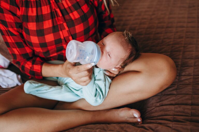 crop anonymous mother feeding adorable newborn baby with bottle while sitting on bed with crossed legs in light bedroom
