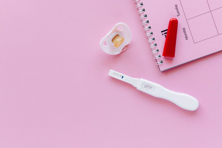 a positive pregnancy test on a pink surface with a pacifier and planner.