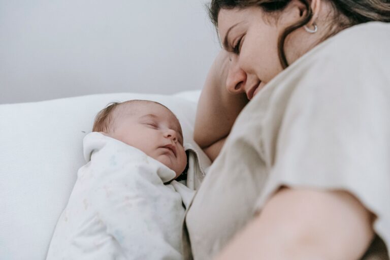 side view of crop positive caring mother looking at sleeping infant baby while lying on comfortable bed in light bedroom