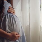 a peaceful moment of a pregnant woman cradling her belly by a sunlit window indoors.