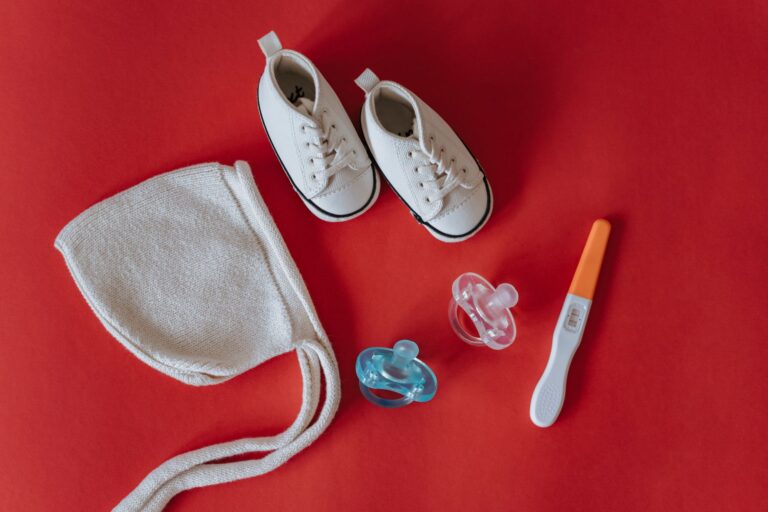 flat lay of baby shoes, pacifiers, hat, and pregnancy test on red surface.