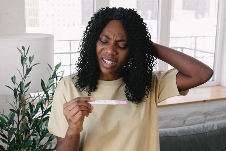 frustrated african american female in casual clothes looking at pregnancy test in hand while standing in light living ro