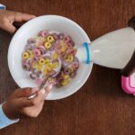 a top view of hands pouring milk into a bowl of colorful cereal on a wooden table.