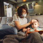 a concerned mother wearing a mask checks her child's temperature on the sofa.