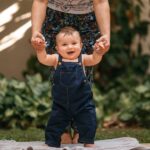 cheerful baby in overalls being supported while taking first steps outdoors.