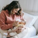 content mother feeding cute infant baby with bottle while sitting on bed with white cushions in light bedroom at home