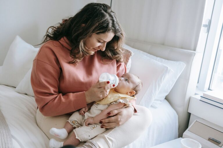 content mother feeding cute infant baby with bottle while sitting on bed with white cushions in light bedroom at home