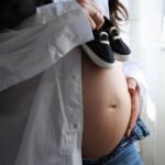 pregnant woman in casual attire holding baby shoes on her belly indoors, symbolizing anticipation.