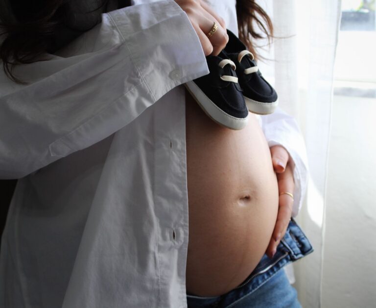 pregnant woman in casual attire holding baby shoes on her belly indoors, symbolizing anticipation.