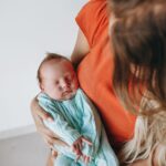 crop anonymous mother with long hair with sleeping baby in arms sitting in light room near white wall at home