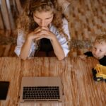 a woman works on a laptop at a wooden table with a child playing with a toy truck beside her.