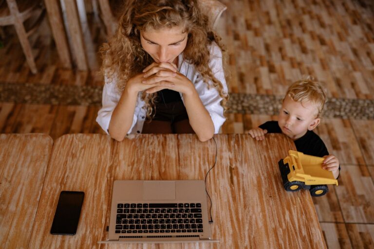 a woman works on a laptop at a wooden table with a child playing with a toy truck beside her.